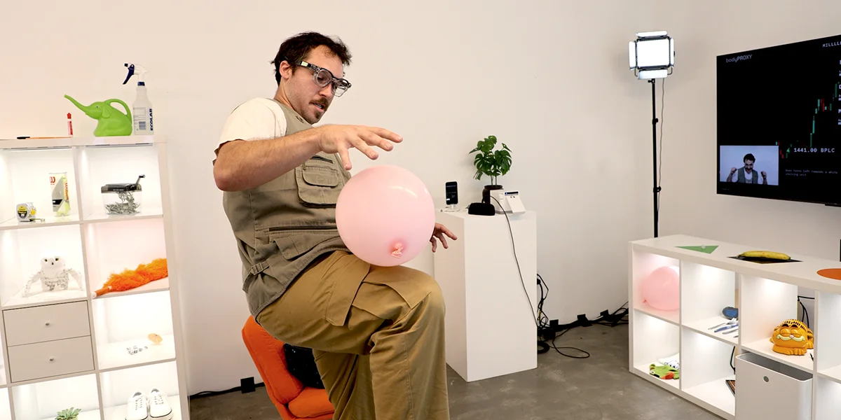 A man in glasses and cargo pants sits on an orange chair, reaching toward a pink balloon in a white room.