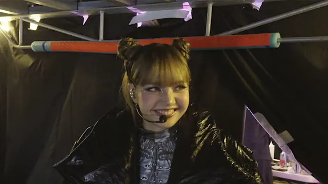 A smiling young girl in a black costume with a headset mic, backstage under metal scaffolding.