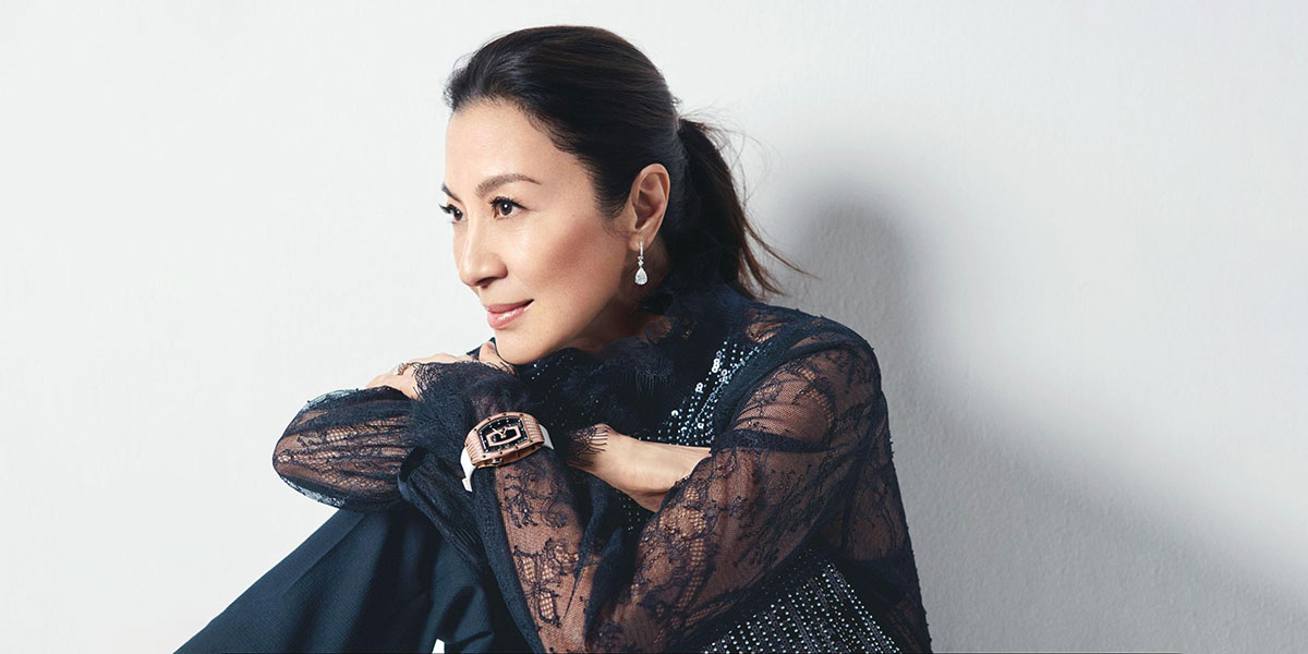 Portrait of Michelloe Yeoh sitting against a white wall with her arms crossed upon her knee