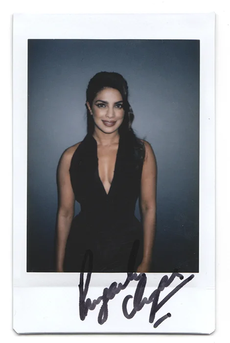 Signed Polaroid photo of a woman in a black deep-V outfit, smiling, against a blue-gray background.