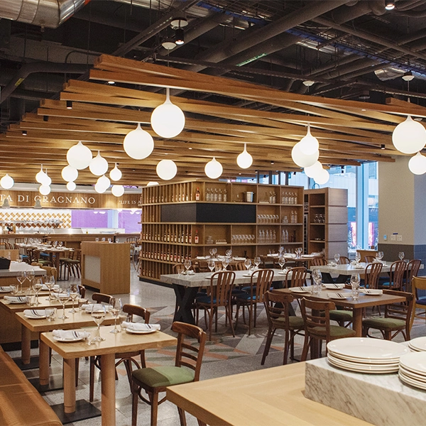 Spacious restaurant dining room filled with wooden tables and chairs, featuring wooden ceiling slats hanging with glowing spherical pendant lights.