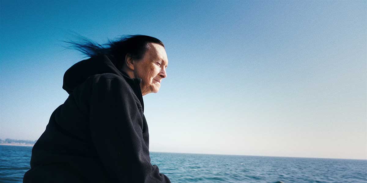 Elderly woman in black hoodie with wind-blown dark hair gazing over a calm blue ocean.