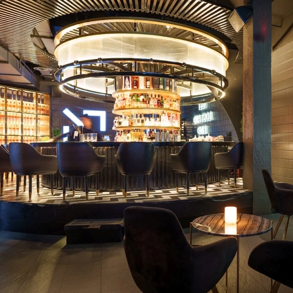 A stylish modern bar with dark swivel chairs surrounding a central circular counter and a glowing bottle display.