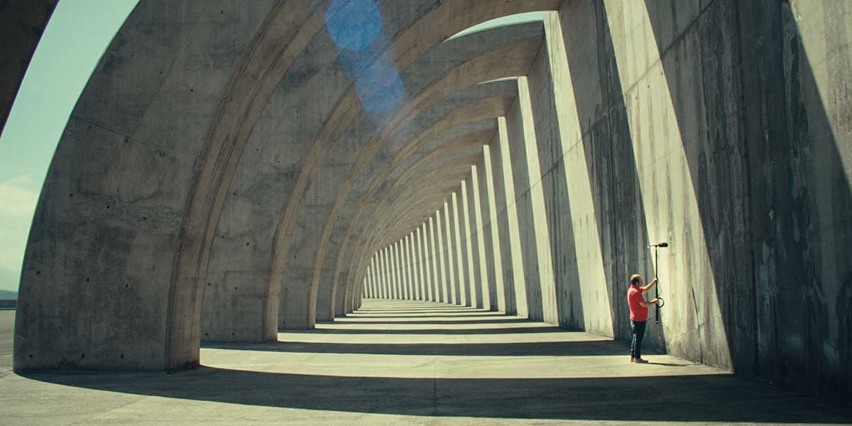 Person in red shirt holding a boom in a concrete archway corridor with dramatic repeating shadows and perspective