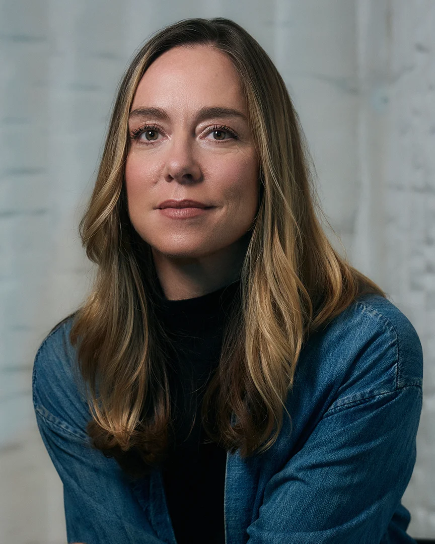 Woman with long brown hair wearing a black turtleneck and denim jacket, looking at camera.