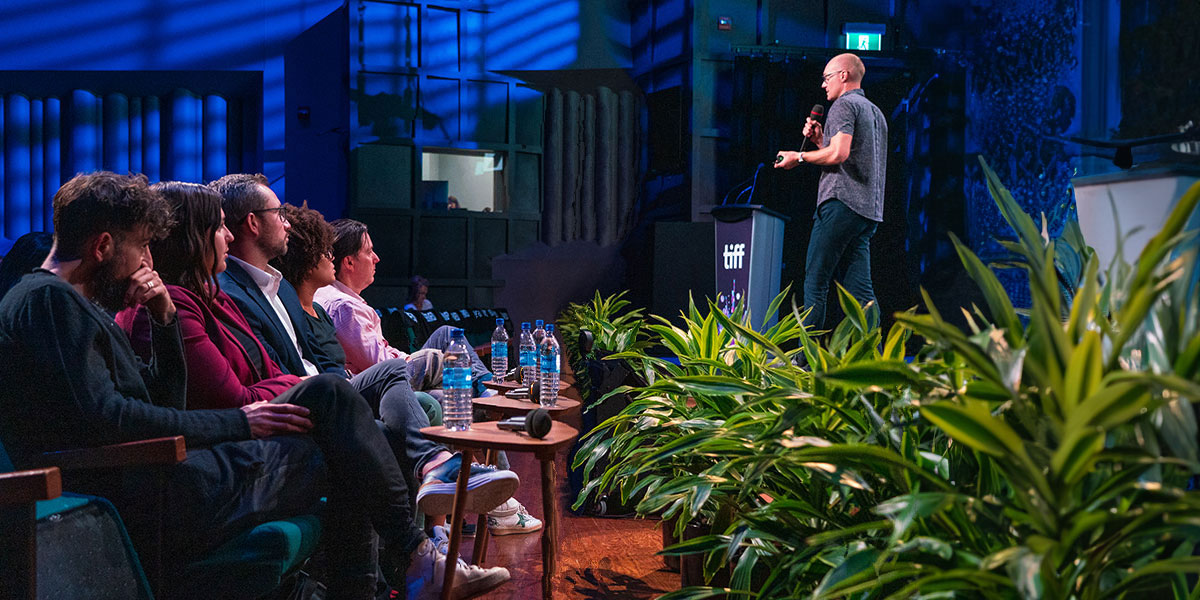 Speaker presenting on stage to audience in blue-lit venue with tropical plants in foreground