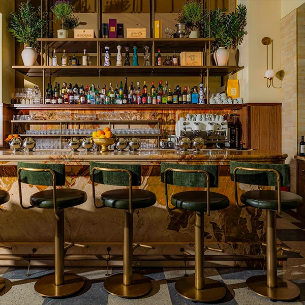 Elegant bar with marble counter, green and gold stools, shelves of liquor bottles, and lush potted plants.