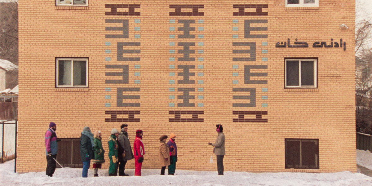 Eight people lining up facing a man in front of an off-orange building in the midst of winter