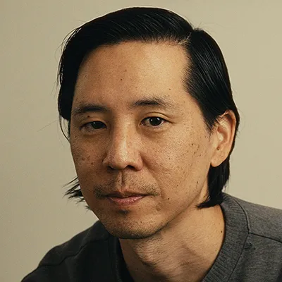 Close-up portrait of an Asian man with black hair wearing a grey t-shirt against a beige background