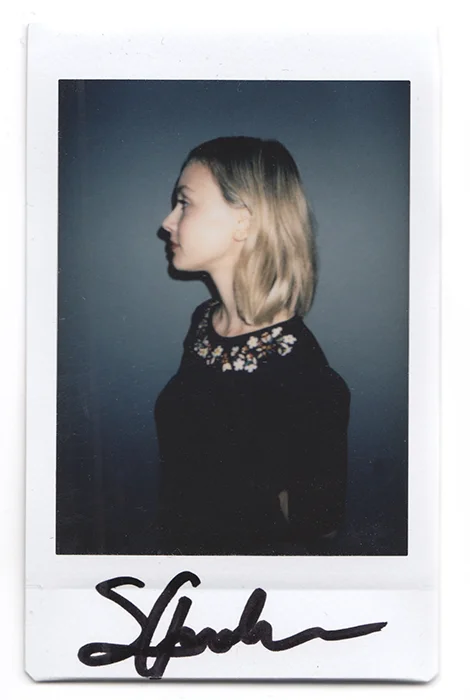Signed Polaroid photo of a young blonde woman in profile, wearing a black floral-collar top, dark blue background.