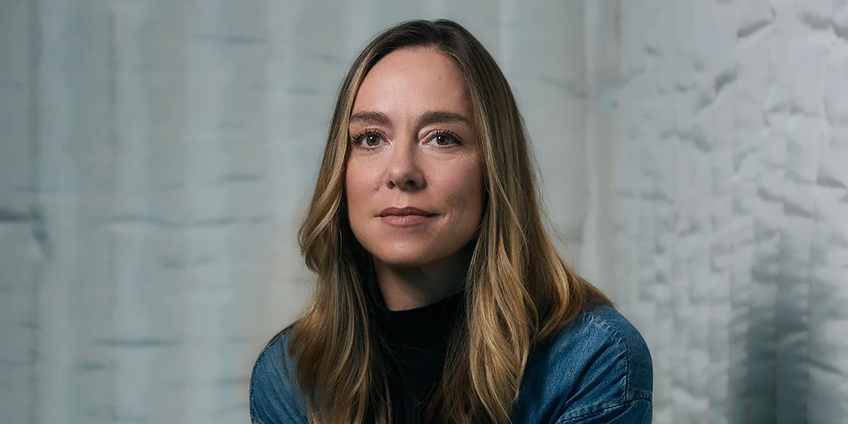 A woman with long brown hair wearing a blue denim jacket over a black turtleneck, posing against a light gray wall.