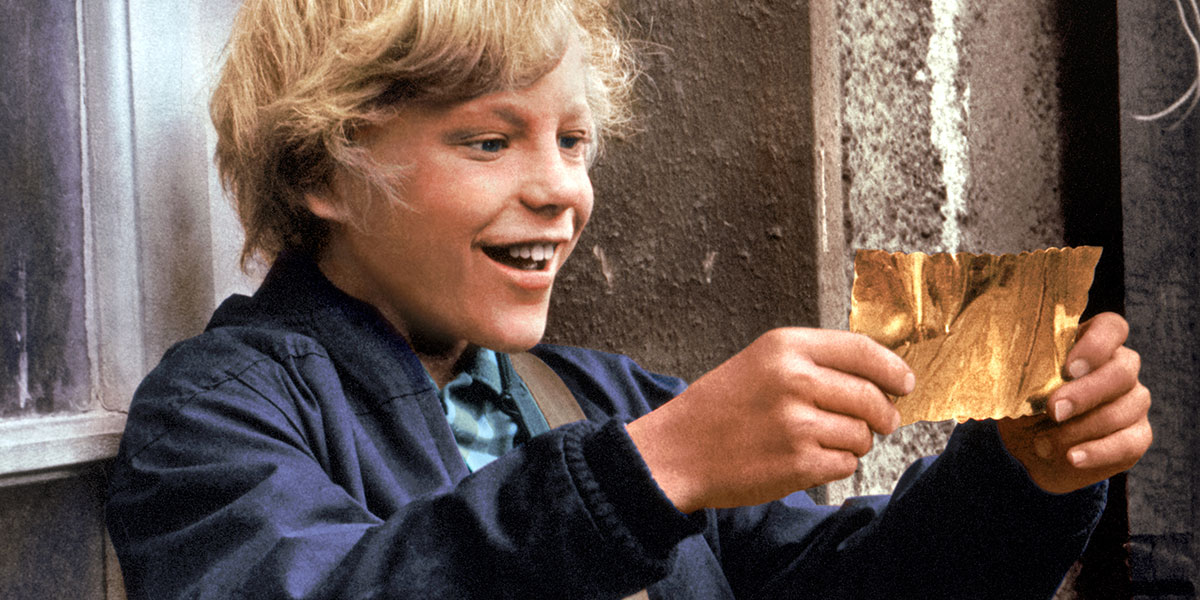 A smiling blond boy in a blue jacket holds a golden ticket with excitement.