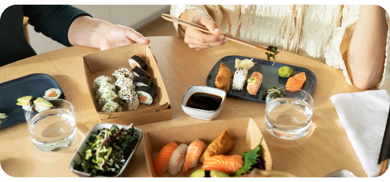 Zwei Personen teilen sich Sushi aus Kraftpapier-Boxen an einem Holztisch, mit Maki, Nigiri, Sojasauce, Algensalat und Wassergläsern