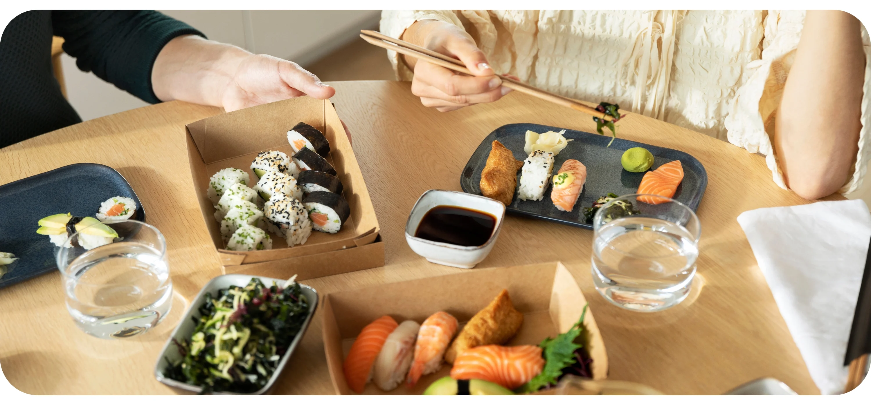 Zwei Personen teilen sich Sushi aus Kraftpapier-Boxen an einem Holztisch, mit Maki, Nigiri, Sojasauce, Algensalat und Wassergläsern