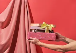 A person’s hands holding a stack of wrapped gift boxes, including one with a yellow ribbon and another with pink confetti paper, set against a soft pink fabric and coral background.