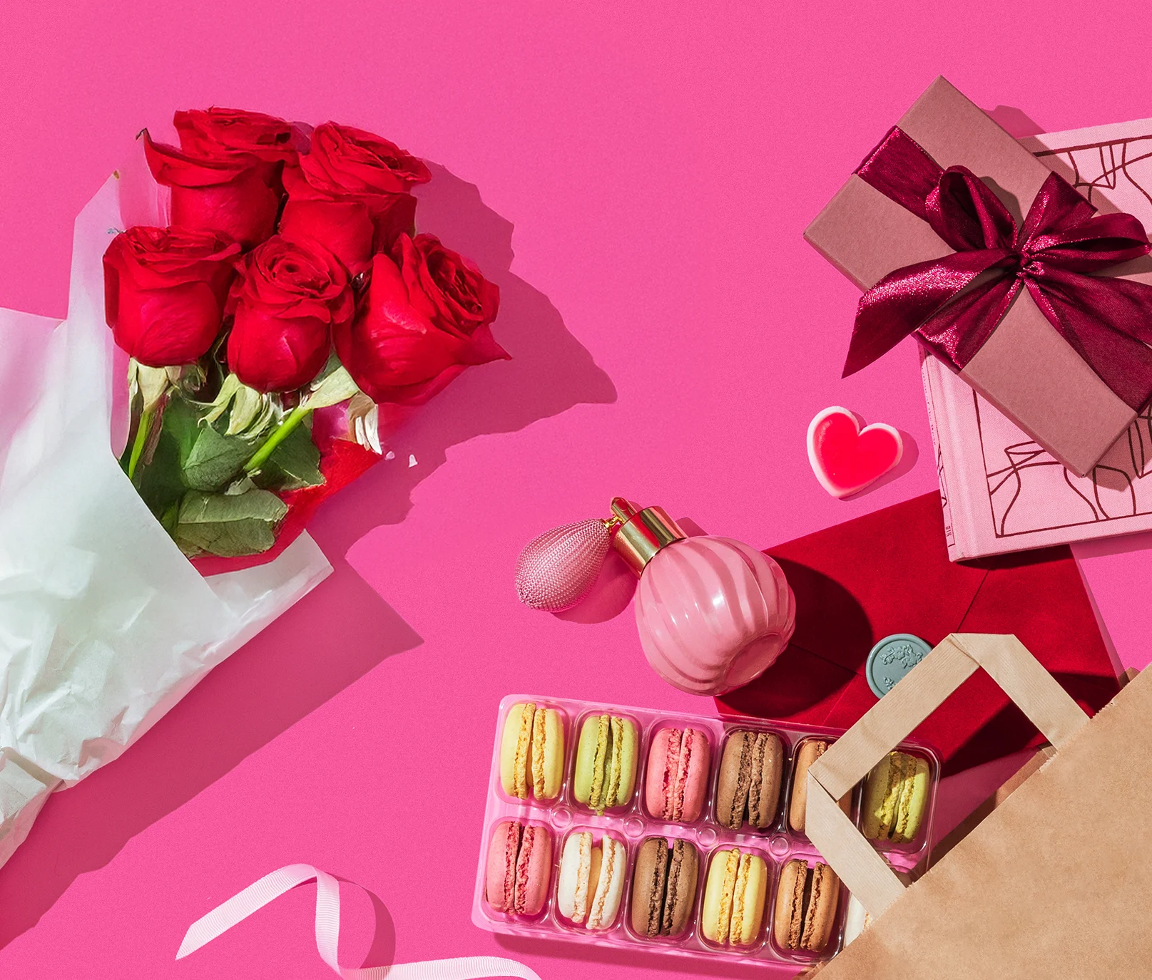 Flat lay of Valentine’s Day gifts on a pink background, including a bouquet of red roses, wrapped gift boxes, a pink perfume bottle, heart-shaped candy, red envelope, and a box of colorful macarons.