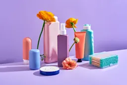 A pastel-toned arrangement of self-care and beauty essentials including bottles, tubes, cotton swabs, and skincare containers, styled with bright orange flowers on a lavender background.