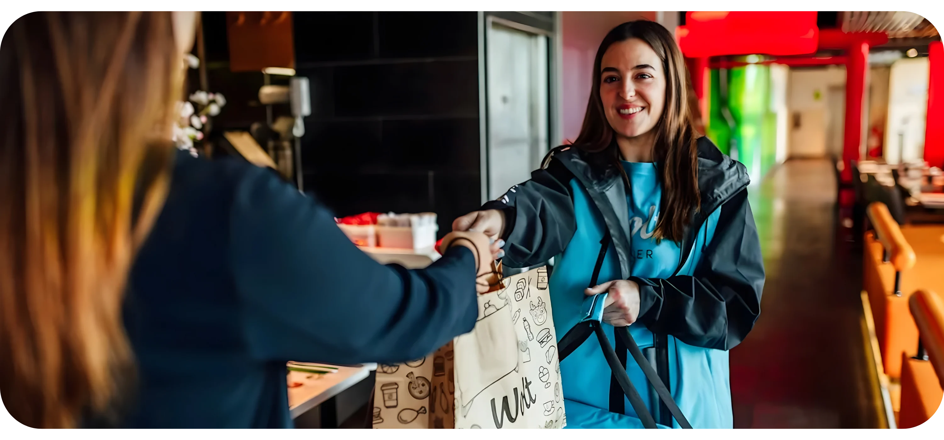 A restaurant employee hands a paper Wolt delivery bag to a courier wearing a blue and black jacket.