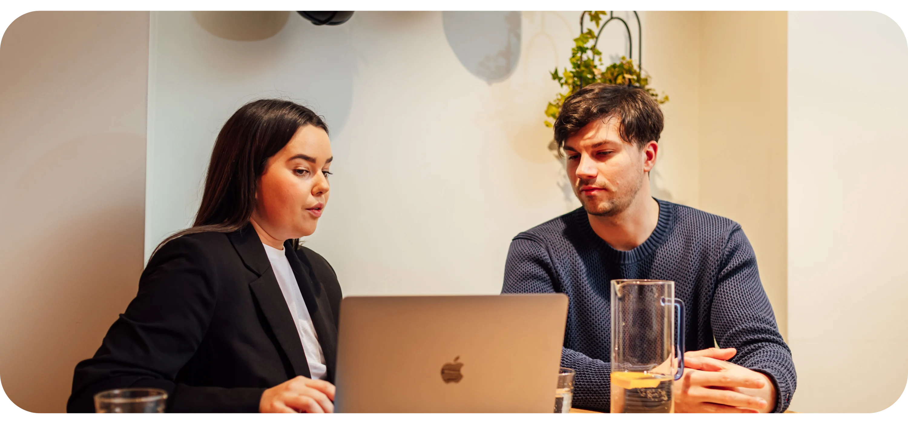 A man and woman sit at a table talking while looking at a laptop, with a water pitcher and glasses nearby.