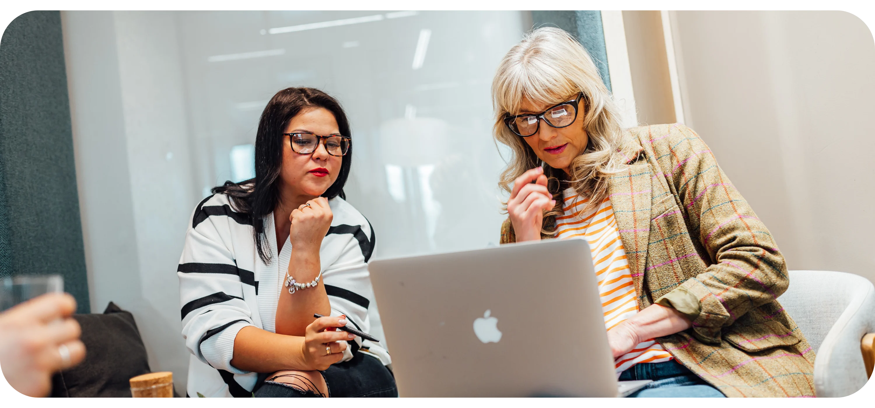Two women sit closely looking at a laptop screen, one wearing glasses and gesturing while talking.