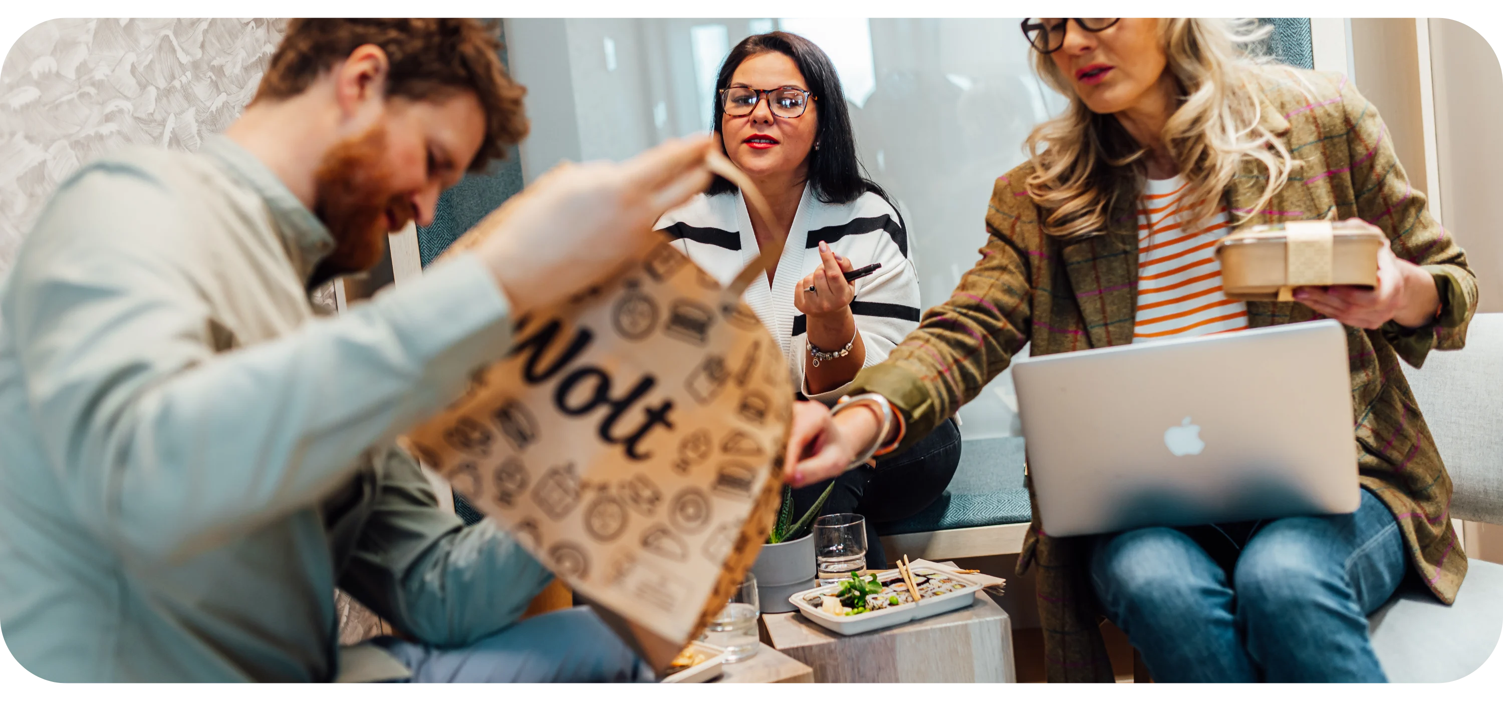 A man holds open a paper Wolt bag while two women look on, one with a laptop and a food container nearby.