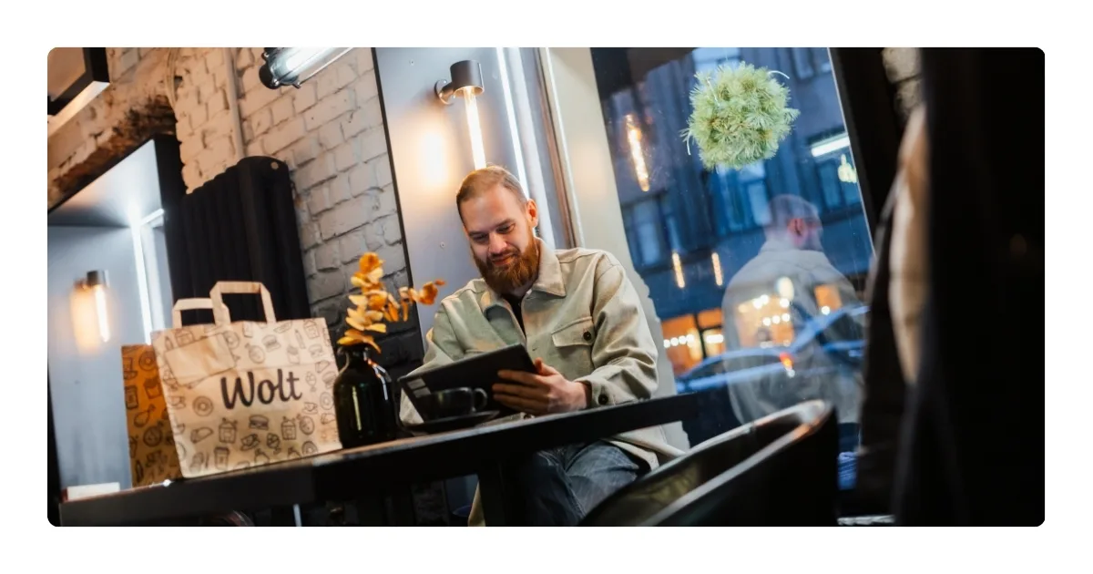 A bearded man in a beige overshirt looks at a tablet at a café table, with a Wolt paper bag, a coffee cup, and a vase of dried flowers beside him