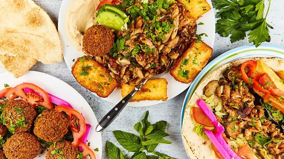 Teller mit Shawarma-Fleisch, Falafel, gebratenem Halloumi, Hummus, Pita-Brot, Tomatenscheiben, eingelegtem Gemüse und Kräutern, dazu ein Wrap auf Fladenbrot