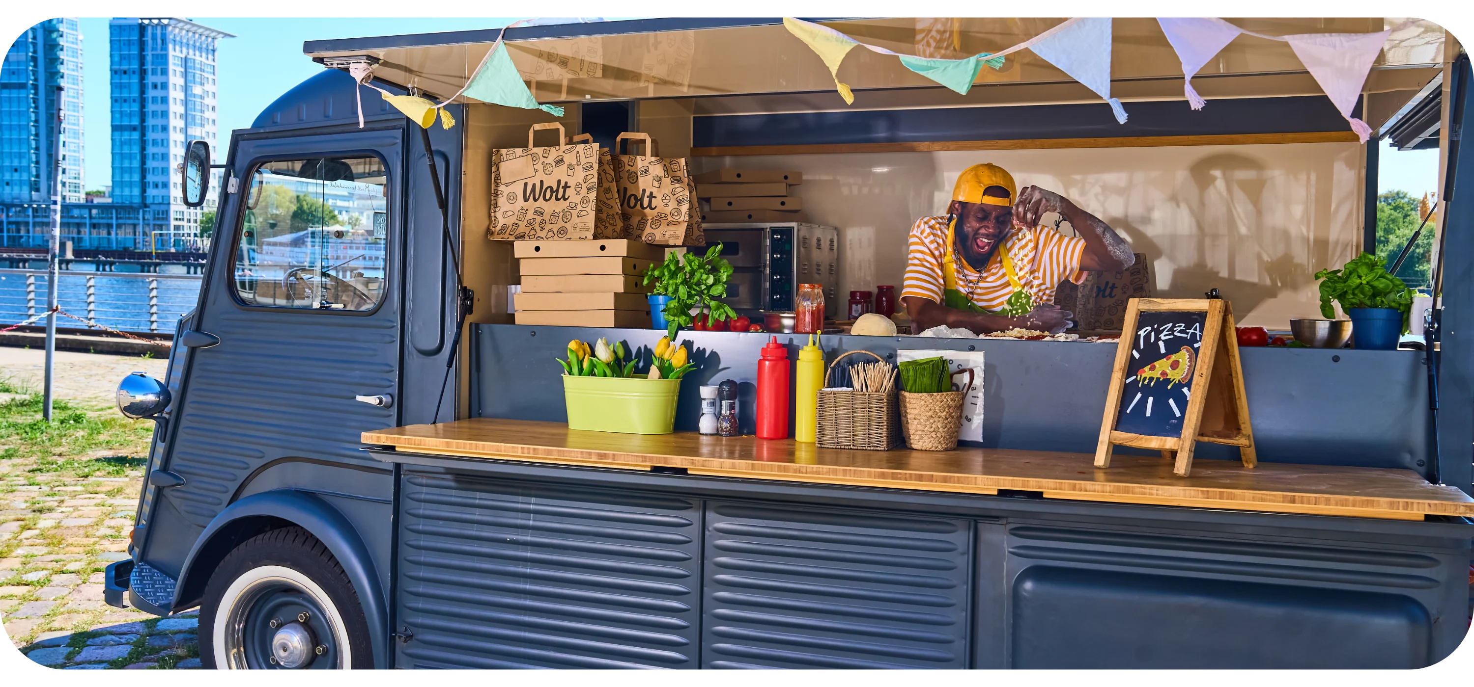 A man laughs while preparing pizza inside a food truck decorated with bunting and paper Wolt bags on the shelf.