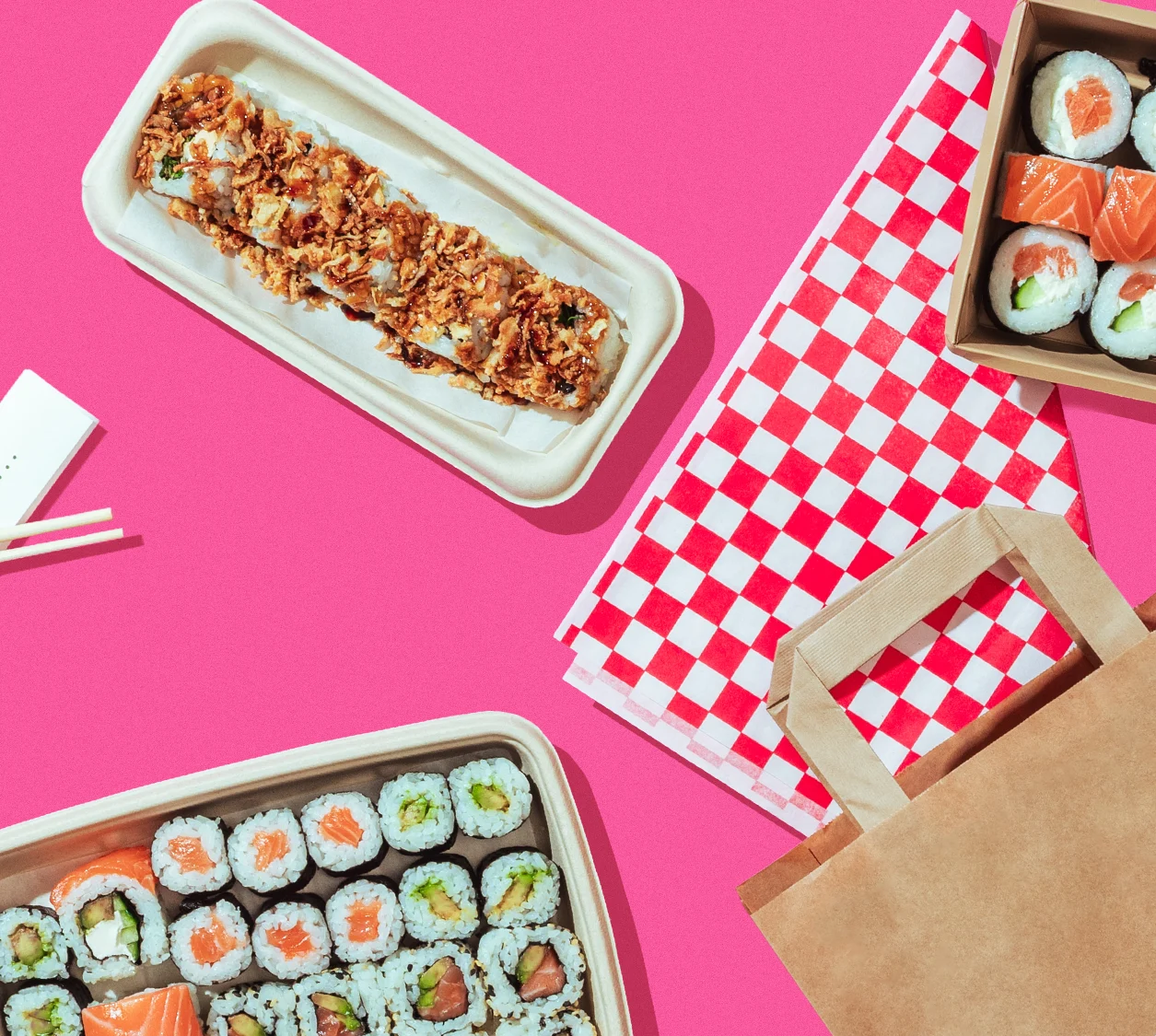 Flat lay of sushi rolls and a crispy sushi roll in takeout containers on a pink background, with a paper bag and checkered napkins.