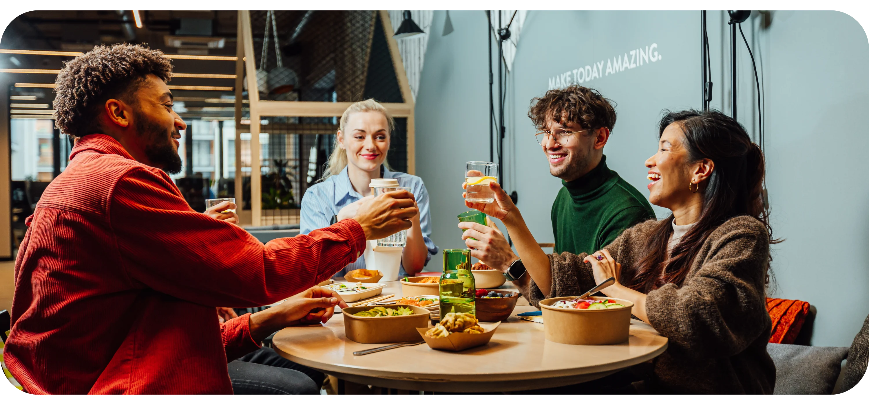 Four people sit around a table with takeaway food containers, smiling and raising their glasses together.