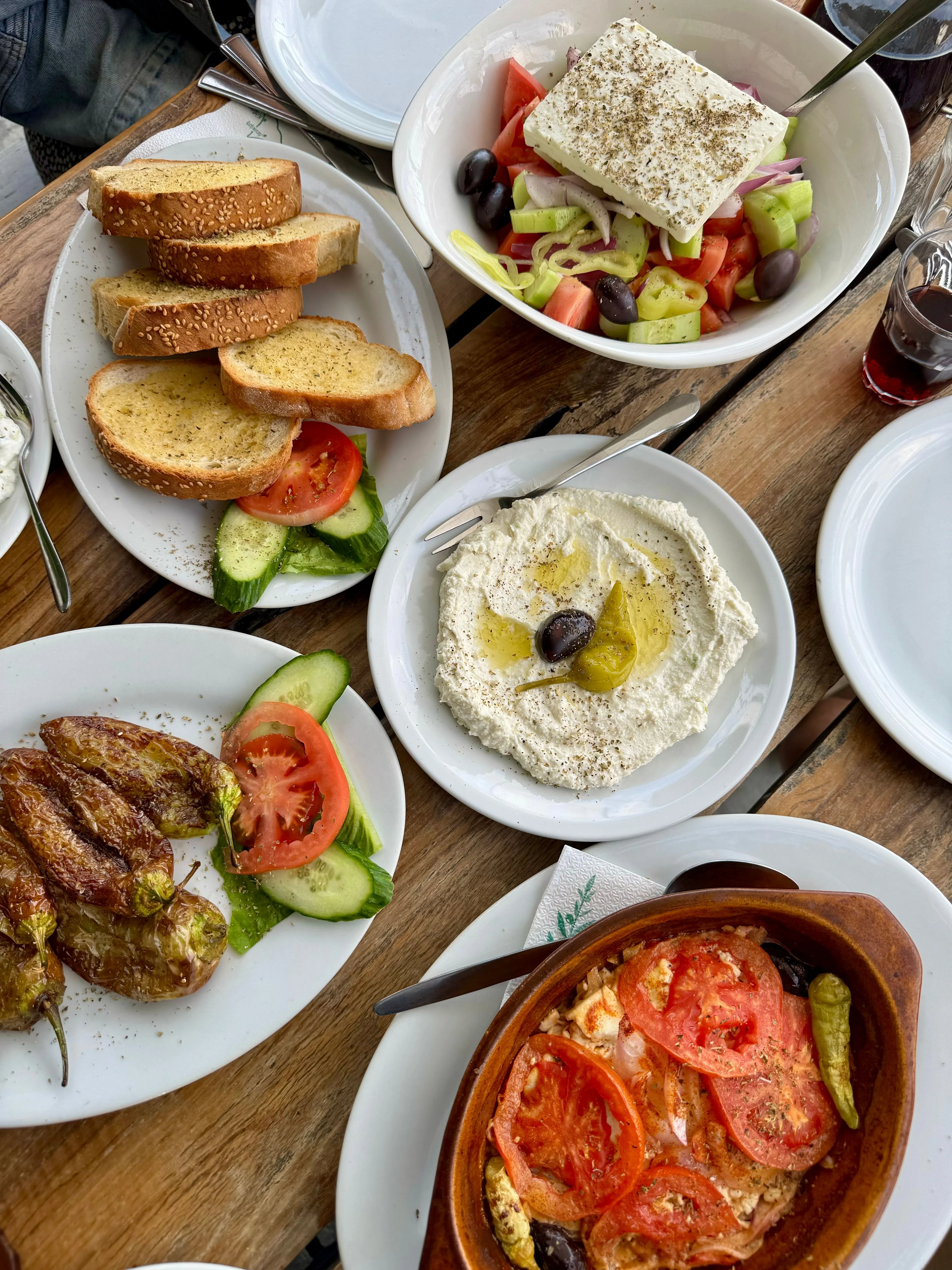 Ein griechisches Gericht-Arrangement auf einem Holztisch, mit griechischem Salat mit einem Stück Feta und Oregano, einem Feta-Dip, gebratenen Paprika, Sesambrot, gebackenem Feta mit Tomate und einem Glas Rotwein