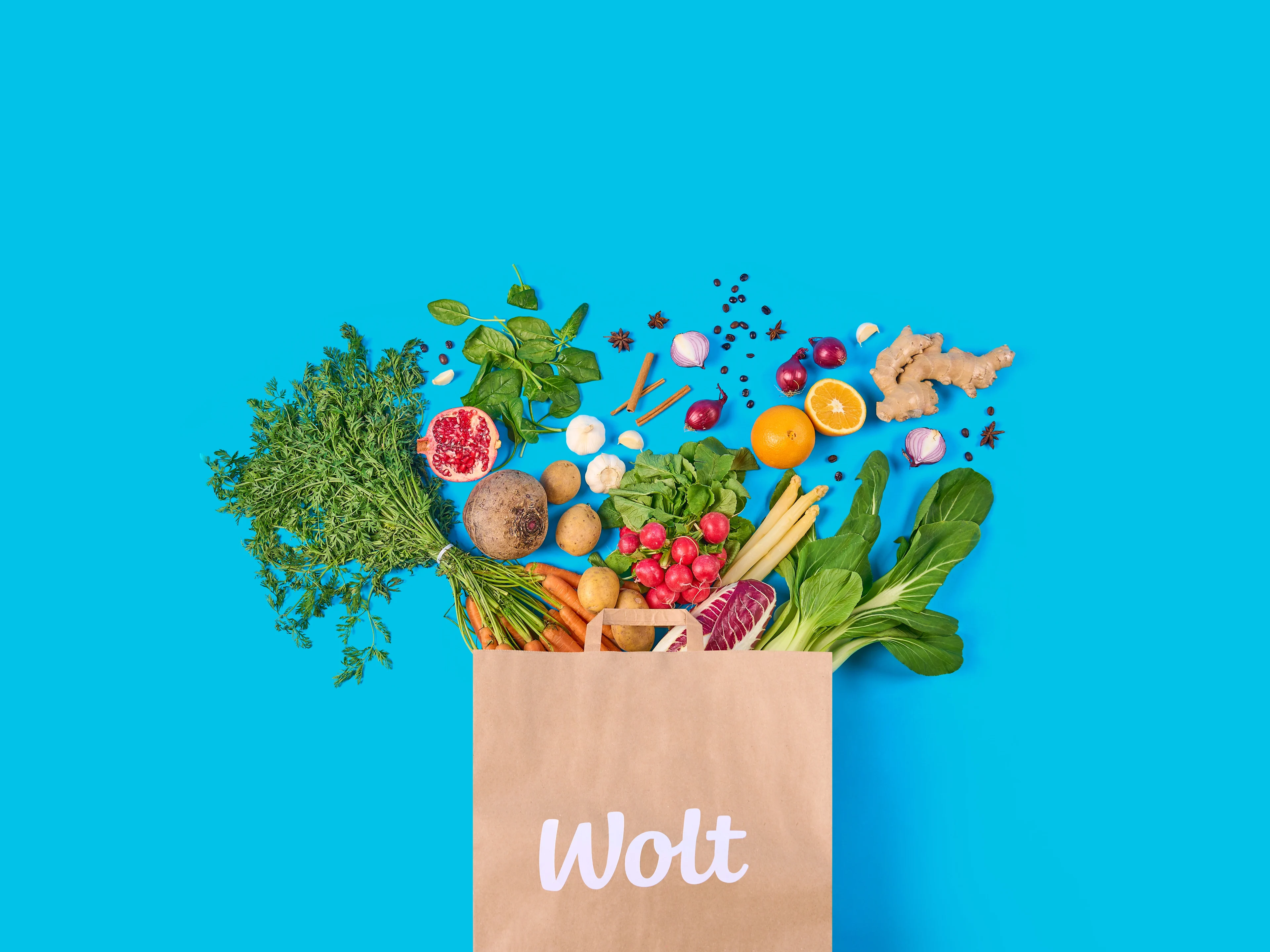 Paper grocery bag with the Wolt logo filled with fresh vegetables, fruits, and herbs spilling out against a bright blue background.