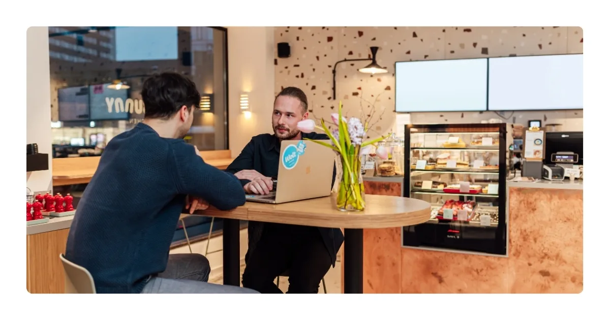 Wolt representative and a café owner discussing business on a laptop at a patisserie counter with pastries and fresh tulips