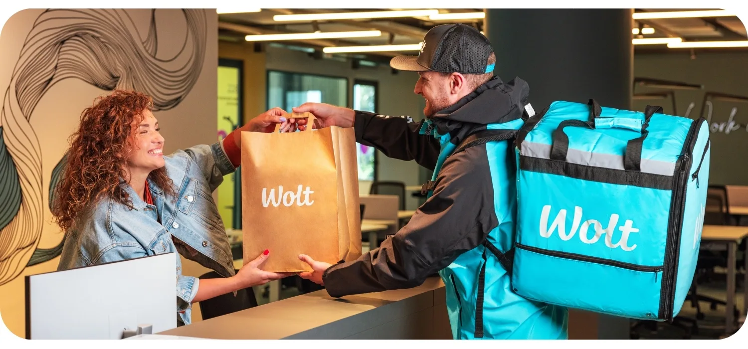 Eine lachende Wolt-Partnerin in Markenkleidung übergibt einem Händler in einem Restaurant eine Wolt-Papiertüte