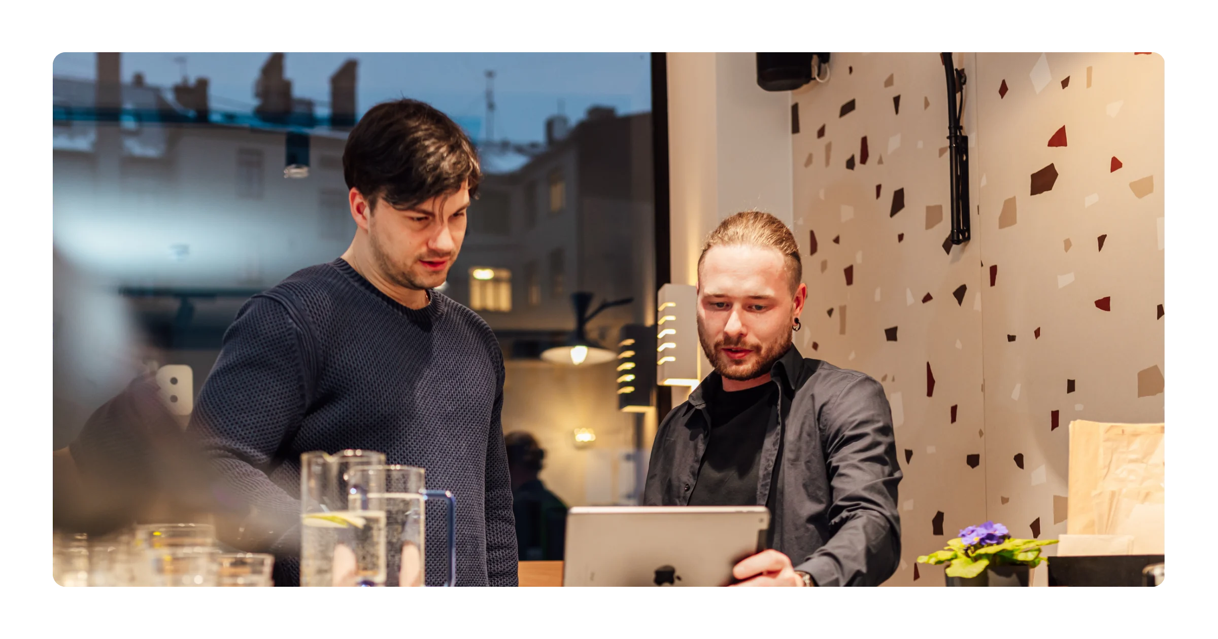 Two men stand at a restaurant counter looking at a tablet screen, with water glasses in the foreground.
