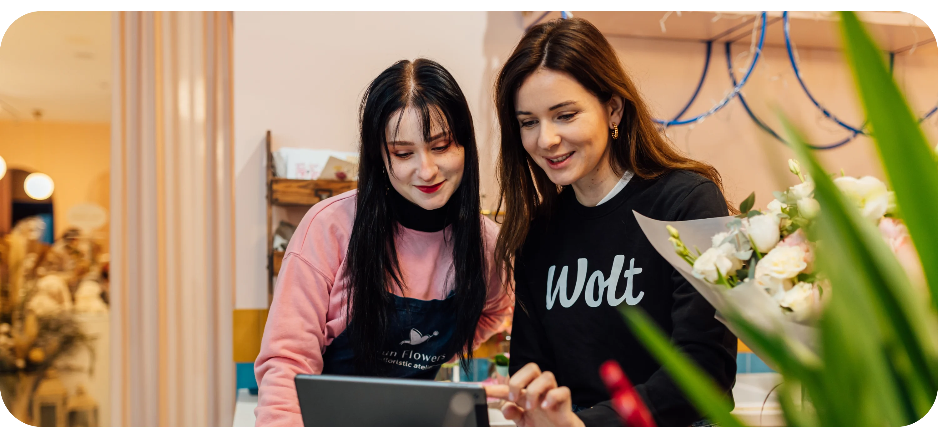 Two women in a flower shop look at a tablet screen, one wearing a black Wolt sweatshirt, bouquet visible.