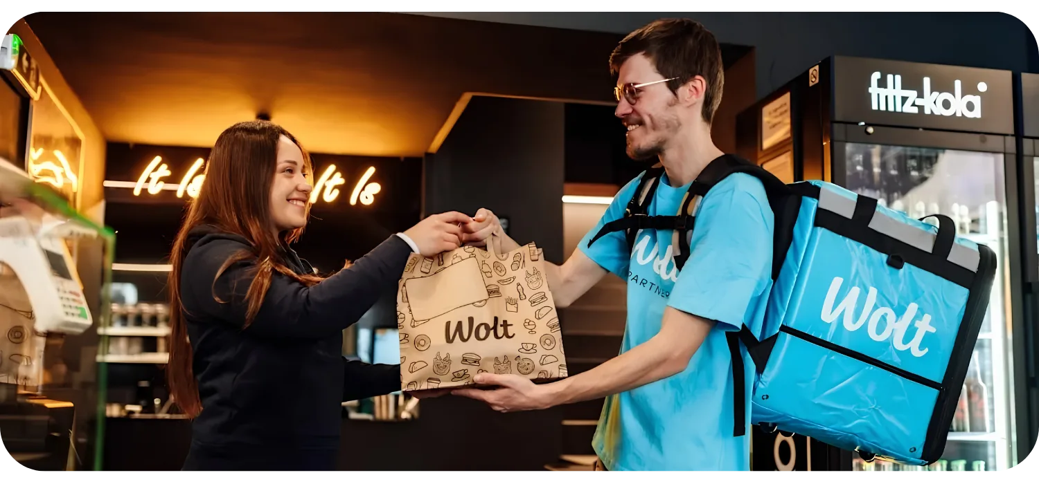 A shop employee hands a packed Wolt order to a courier wearing a blue Wolt delivery backpack at the counter