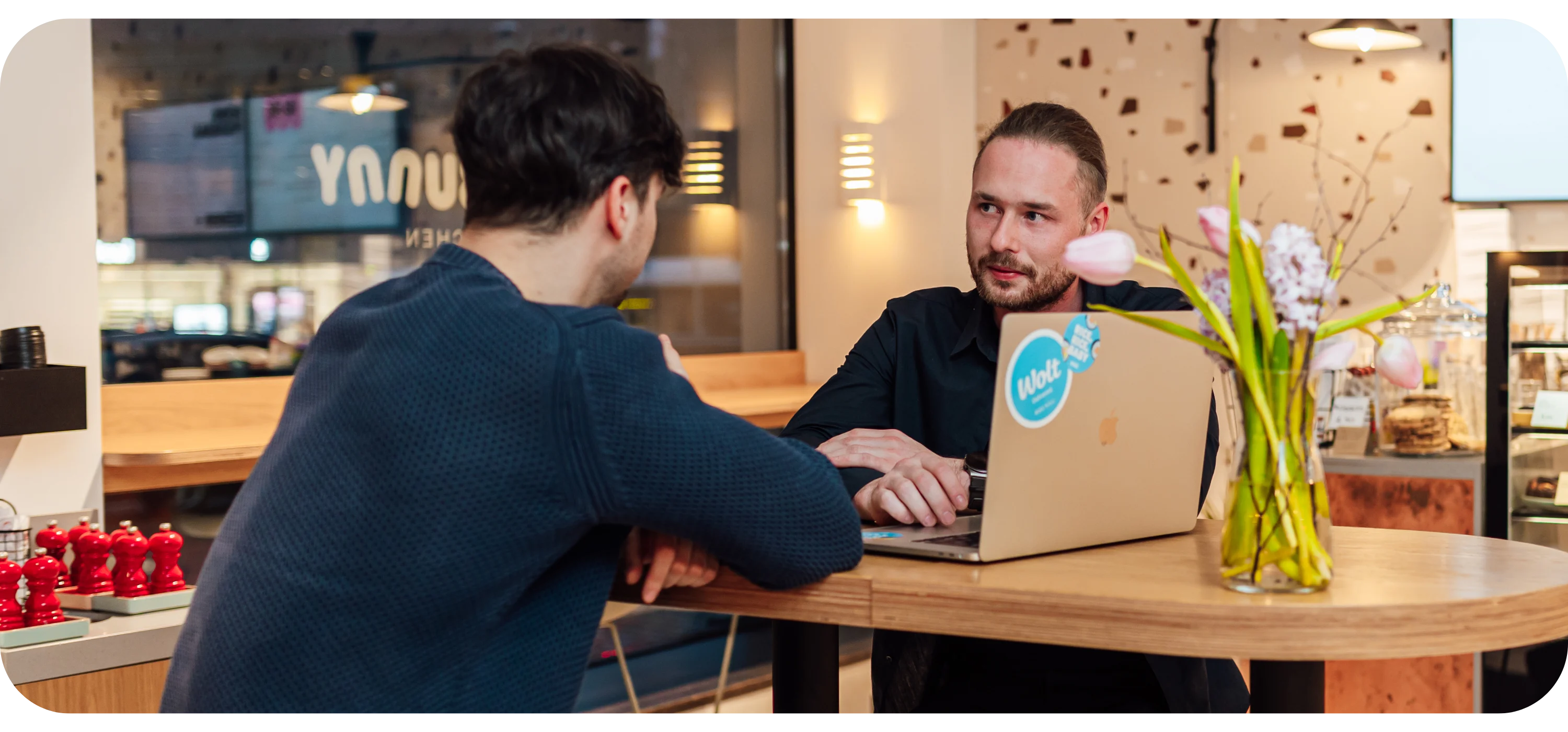 Two men sit at a wooden table talking, one with a laptop featuring a Wolt sticker, a vase of flowers nearby.