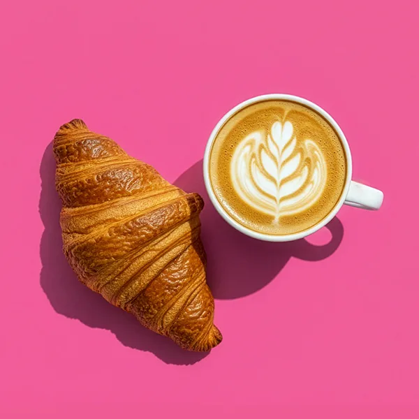 Croissant served beside a latte with leaf-shaped foam art on a pink background.