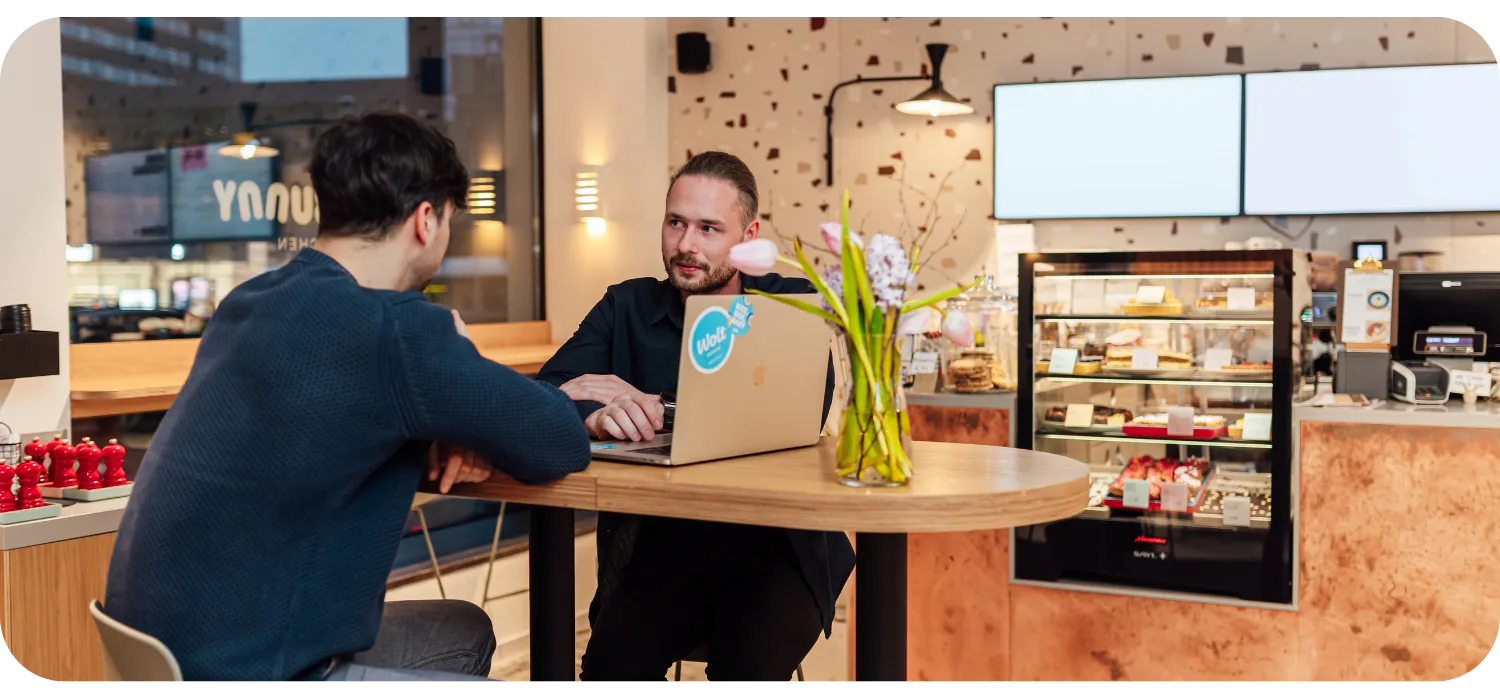 A café owner with a Wolt-stickered laptop talks with a partner at a table, with a pastry display in the background