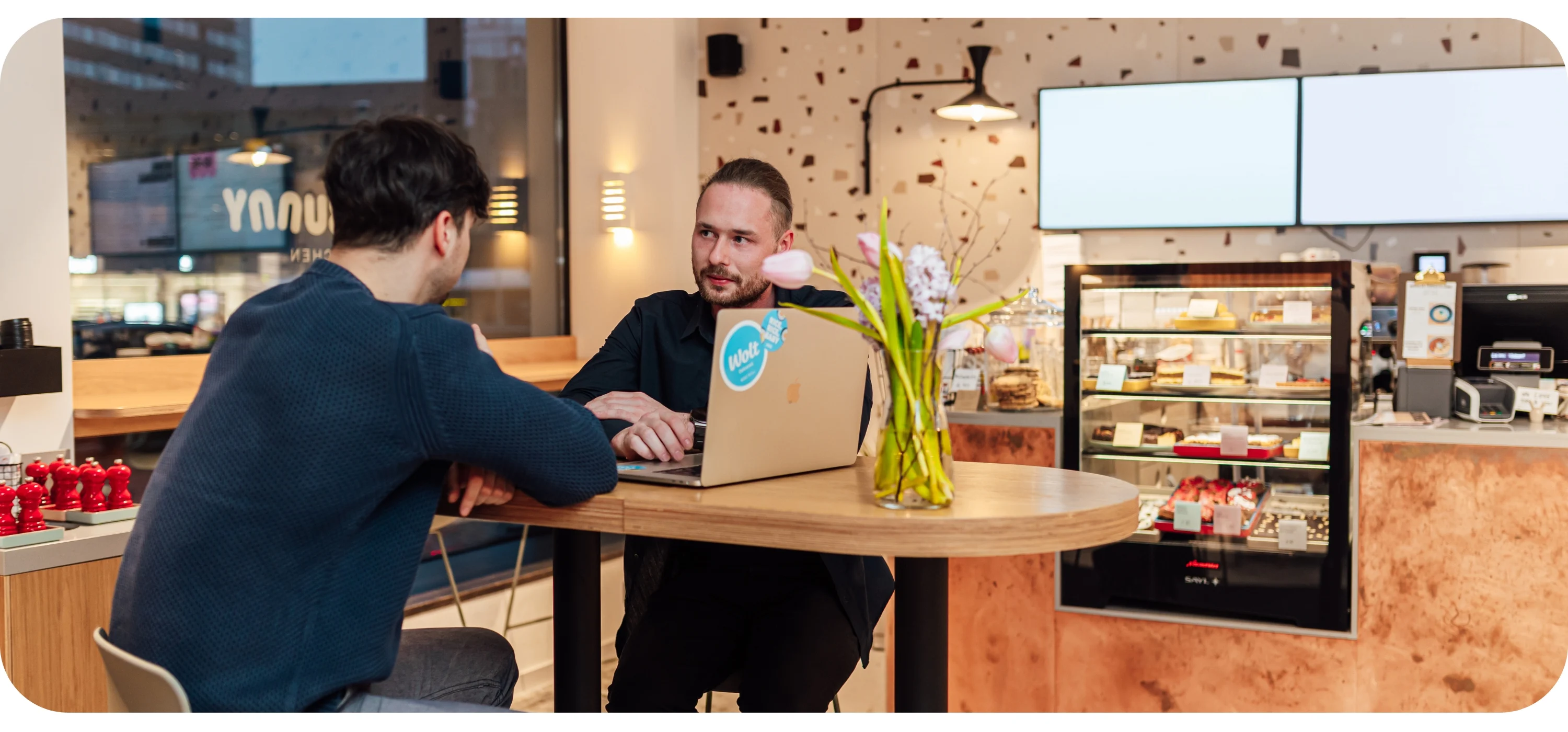 A café owner with a Wolt-stickered laptop talks with a partner at a table, with a pastry display in the background