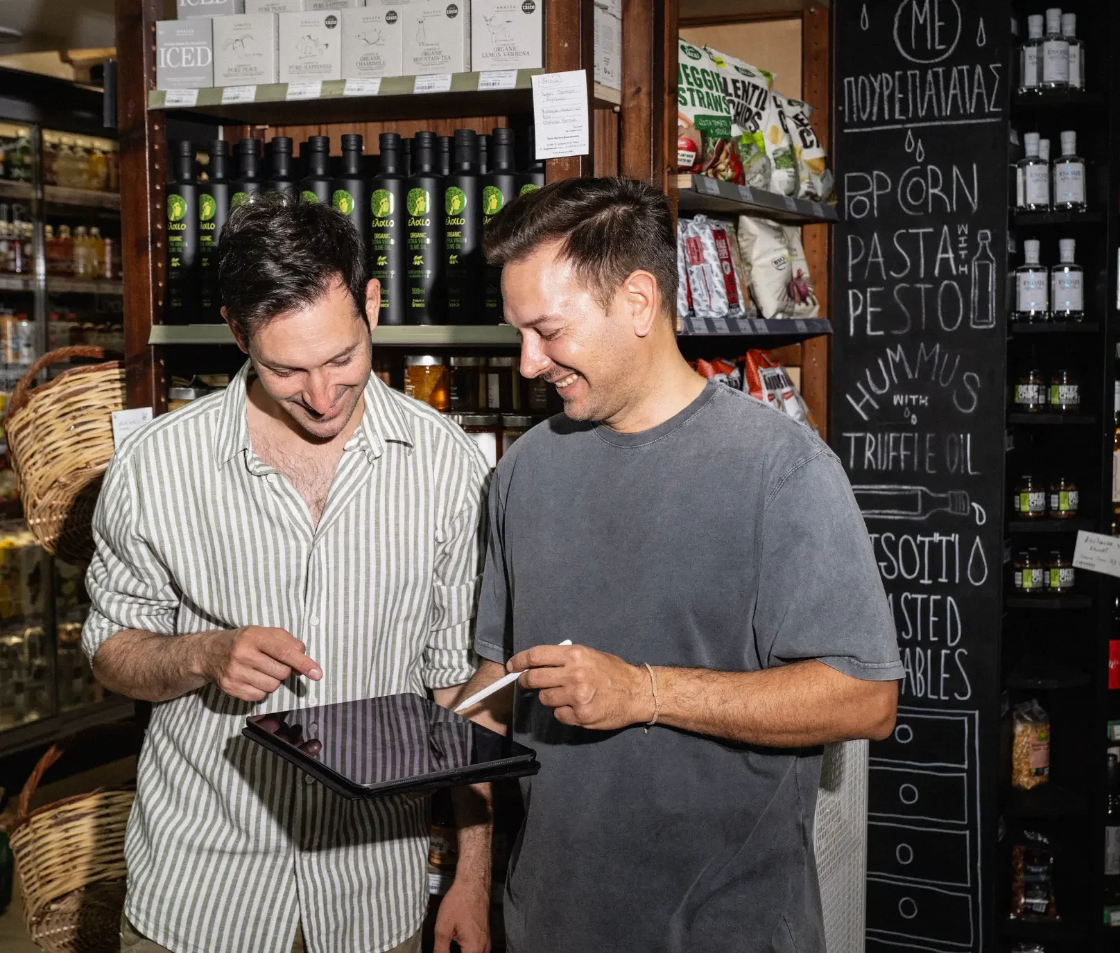 Two brothers inside their 4 Seasons Bio grocery store in Athens smiling as they look at a tablet together, checking orders among shelves of organic products and olive oil bottles.