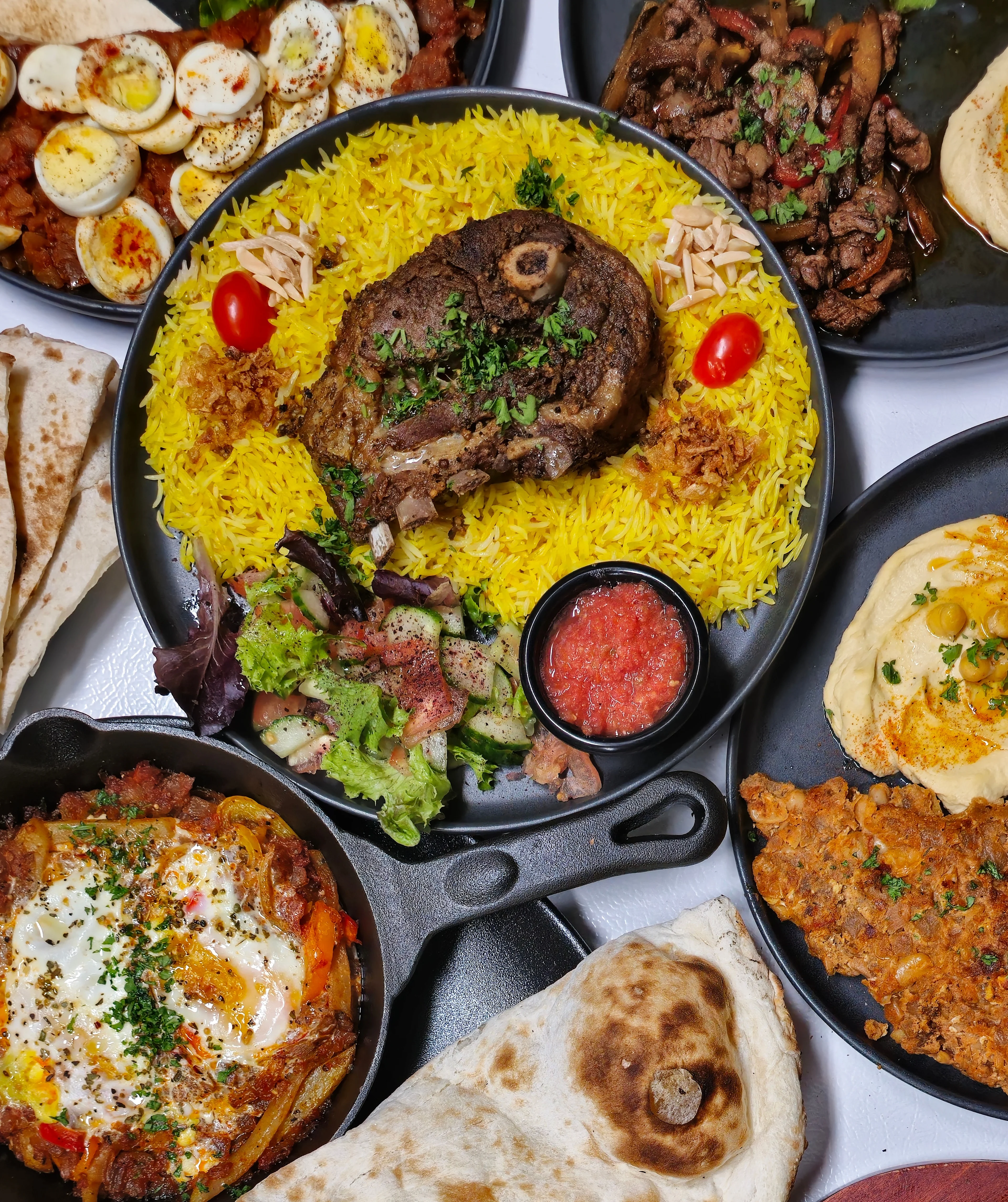 Ein orientalisches Gericht-Arrangement mit Lammhaxe auf Safranreis, Hummus, Shakshuka, gebratenem Rindfleisch mit Paprika, Scheiben von gekochten Eiern in Sauce, Fladenbrot, Salat und Tomaten-Dip