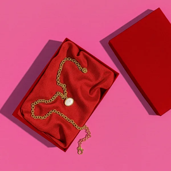 Gold necklace displayed on red fabric inside an open jewelry box on a pink background.