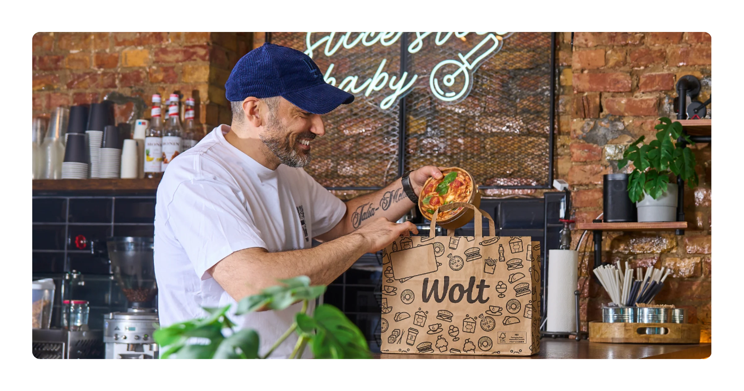 A smiling man in a cap places a bowl of food into a paper Wolt bag on a café counter with a neon sign behind him.