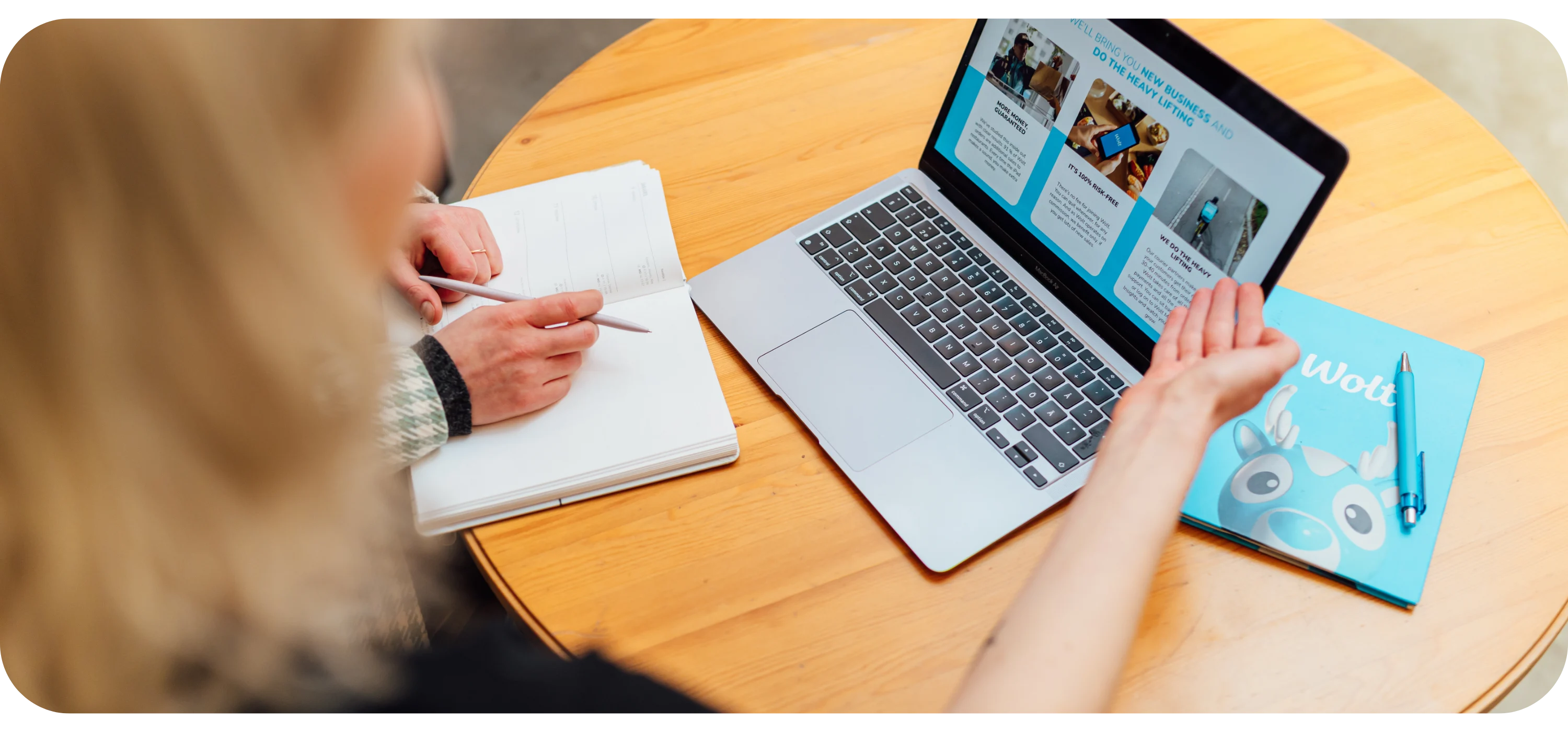 A person writes in a notebook beside a laptop showing a Wolt web page, with a blue Wolt notebook nearby.