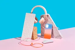 A minimalist setup featuring pastel-colored tech accessories — wireless headphones, a white keyboard, a smartphone with a blue and orange gradient case, a computer mouse, and cables — arranged on a pink and blue backdrop with marble and stone props.