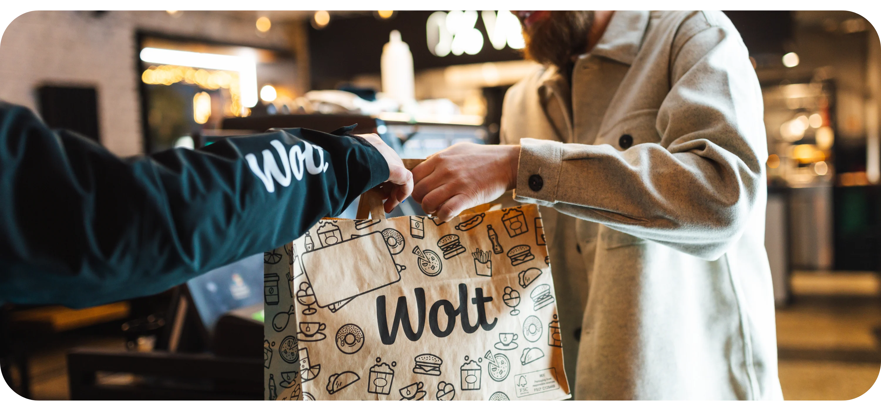 A person in a Wolt sleeve hands a paper delivery bag to another person's outstretched hand.