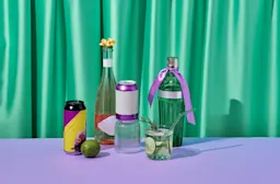 A stylish arrangement of drinks including bottles with ribbons, cans, and a glass of cocktail garnished with lime and rosemary, set on a lavender surface with a green satin curtain backdrop.