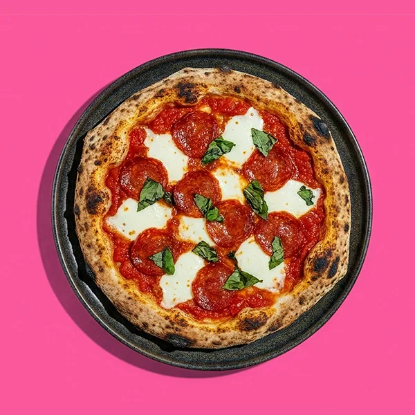 Pepperoni pizza with melted mozzarella and basil leaves on a pink background.